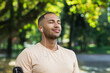 © Liubomir - Close-up portrait of sportsman in park, hispanic man jogging in park with eyes closed breathing fresh air and resting, jogging with headphones listening to music and online radio and podcasts.