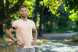 Portrait of successful man in park hispanic man with headphones listening to music and audiobooks online podcasts, smiling and looking at camera while jogging and doing fitness exercise.
