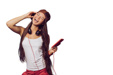 Charming young brunette girl with long hair dressed in white t-shirt, baseball cap and red pants, listening music with phone, headphones on her head, shows rock hand gesture over transparent backdrop.