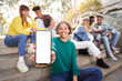 Smily girl showing smartphone with empty screen at street city with friends. Carefree young millennial people in stylish casual outfit having fun together outdoors background. Mockup copy space banner