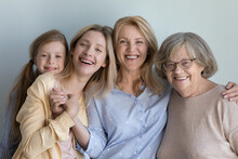 Happy Little Child, Cheerful Young Mom, Middle Aged Granny, Elderly Great Grandma Portrait. Four Girls And Women Of Different Generations Standing At Grey Wall Background, Hugging With Love, Smiling