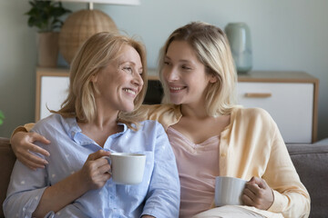 Positive caring young adult daughter woman and happy middle aged mom sitting close together on couch, talking over mug of tea, laughing, enjoying warm relationships, family meeting