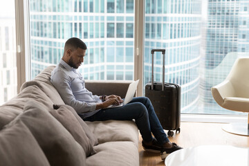 Serious busy African businessman or traveler working on laptop, texting message, sending corporate e-mail sits on couch in luxury skyscraper hotel room on business trip. Lifestyles, modern technology