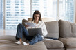 © fizkes - Busy woman sitting on couch with laptop on laps holding bills, makes easy remote e-payment through e-banking application, student prepare for college exams, studying at home using internet connection