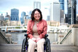 © Austockphoto - Smiling woman with a disability sitting in a wheel chair with tall buildings behind her