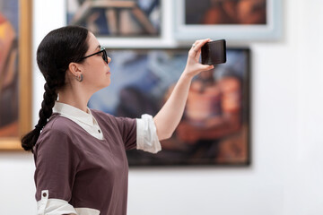 Wall Mural - Cultural education and modern exhibition in gallery. Side view of woman visiting excursion in museum. Student using smartphone and take photo. Concept of Museum's day