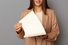 Horizontal Shot Of Faceless Unknown Woman Wearing Beige Official Style Jacket Standing Isolated Over Gray Background, Holding Organizer, Turning Page Of Notepad.