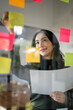 Young smiley attractive businesswoman using sticky notes in glass wall to writing strategy business plan to development grow to success