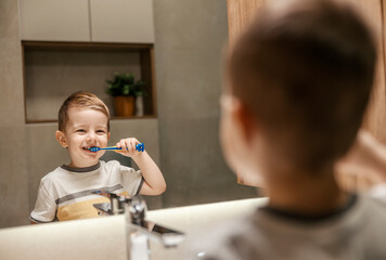 A little boy is standing in front of the mirror and brushing his teeth in the bathroom.