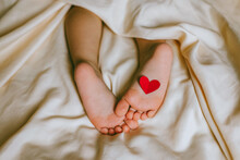 Children's Feet And Heels With Heart On A White Bed To Christmas, Valentine's Day, Mother's Day. Infant Baby Is Sleeping In His Crib. Importance Of Sleep For Babies. Sleep Mode Babies 3 Y.o.
