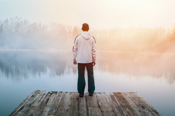 young man in hoodie, hat and pants standing on wooden pier on pond shore with melancholy fog at sunr