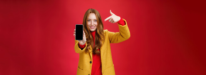 Wall Mural - Pick this phone you never regret. Portrait of friendly-looking attractive and confident redhead glamour girlfriend in yellow coat showing smartphone pointing at mobile as smiling broadly at camera