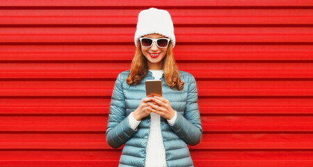 Wall Mural - Portrait of smiling young woman with smartphone wearing jacket, white hat on red background