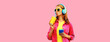Portrait of stylish young woman listening to music in headphones with cup of fresh juice wearing jacket on pink background