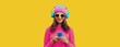 Leinwandbild Motiv Portrait of modern young woman in wireless headphones listening to music with smartphone wearing knitted sweater, pink hat on yellow background