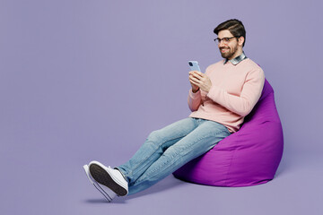 Wall Mural - Full body young happy caucasian IT man he wear casual clothes pink sweater glasses sit in bag chair hold in hand use mobile cell phone isolated on plain pastel light purple background studio portrait.