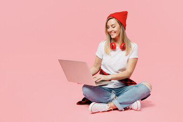 Wall Mural - Full body smiling happy smart young IT woman wear white t-shirt red hat sit hold use work on laptop pc computer isolated on plain pastel light pink background studio portrait People lifestyle concept