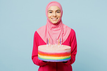 Young happy arabian muslim woman wear pink abaya hijab hold in hand colorful cake with candles isolated on plain pastel light blue cyan background studio portrait. People uae islam religious concept.