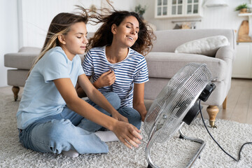 Mother and daughter cooling down near electric fan blowing fresh air, spending time together at home on hot summer days, mom and child sitting on floor in front of ventilator cooler during summer heat