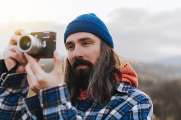 Portrait of attractive traveler man with long hair in a blue beanie against the backdrop of picturesque hills. Photographer traveler taking pictures of sunset on old camera.