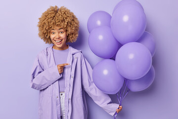 Wall Mural - Festive occasion concept. Positive curly haired woman wears raincoat points at bunch of inflated balloons celebrates bithday isolated over purple background. Female model enjoys celebration.