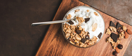 Granola cereal oatmeal with white yogurt, chocolate, fruist and nuts in a bowl on dark wooden board, top view