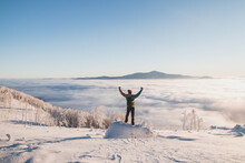 Excited Boy In Winter Clothes Standing On A Stump Celebrating His Successful Summit Of The Mountain. Enjoying The Feeling Of Being Above The Clouds And The Sunrise During A Winter Fairy Tale