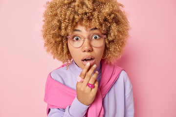 Wall Mural - Image of shocked curly haired woman keeps hand on lips keeps mouth opened stares speechless at camera realizes something amazing wears transparent eyeglasses and pullover isolated over pink background