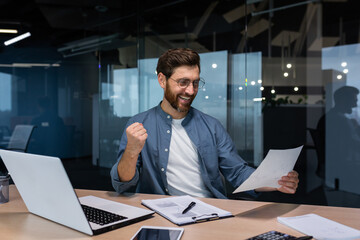 A young man, a businessman, a freelancer sits in the office at the table, holds a document, paper in his hands. He is happy, he points with his hand yes. I received good news, a successful deal