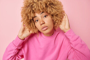 Wall Mural - Headshot of beautiful woman with curly bushy hair keeps hands on head looks directly at camera has calm expression clean healthy skin wears pullover isolated on pink background. Natural beauty concept