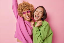 Horizontal Shot Of Two Women Express Positivity And Joy Laugh Happily And Exclaim Loudly Stand Closely Dressed In Casual Jumpers Stand Pleased Isolated Over Pink Background. Friendship And Emotions