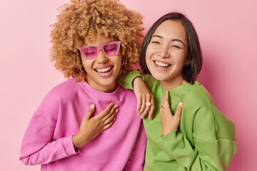 Wall Mural - Overjoyed multiracial women laugh happily smile toothily keep hands on chest have upbeat mood wear sunglasses casual jumpers stand closely to each other isolated on pink background. Positive emotions