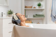 © deliris - Attractive middle aged woman lying in bathtub relaxed listening to music or podcast with headphones smiling with closed eyes. Body-care anti-age recreation skin care and beauty cosmetic concept.