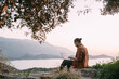 © Anna - A young Caucasian man remotely works with a laptop in a garden on a mountain overlooking the sea and sunset.