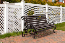 Empty Dark Wooden Bench On The Background Of A White Plastic Fence In A Rural Village. Comfortable Resting Place Under The Open Sky. Close-up, Copy Space.