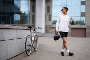 Wall Mural - Smiling african american woman holding helmet and cellphone near bike on street 