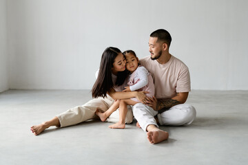 Wall Mural - Young asian parents hugging toddler child together while sitting on grey background