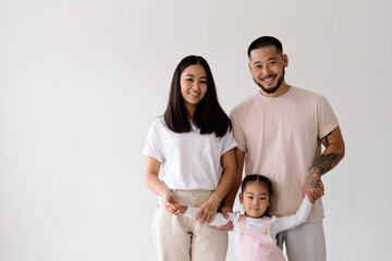 Wall Mural - Smiling young asian parents holding hands of toddler daughter and looking at camera isolated on grey 