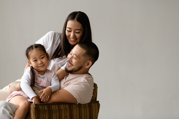 Wall Mural - Happy asian parents looking at toddler daughter while sitting on armchair isolated on grey background 