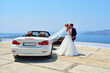 Beautiful young couple bride and groom posing near white cabriolet on Santorini island