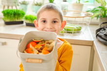 Smiling Boy Holding Compost Bucket Of Orange Peels At Home