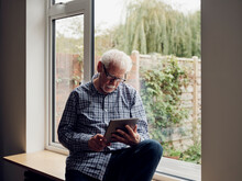 Senior Man By The Window At Home Using Digital Tablet