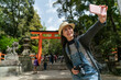 © PR Image Factory - cheerful Asian Japanese girl using mobile phone to take selfie picture with scarlet torii gate at background while traveling to kasuga grand shrine in nara japan on a clear sunny day