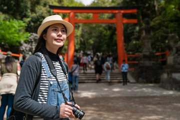 Wall Mural - cheerful Asian Chinese girl traveler carrying camera and looking into distance while admiring beautiful view near a scarlet torii gate of kasuga grand shrine in nara japan