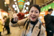 © PR Image Factory - closeup with selective focus of red grilled marinated baby octopus shown to camera by a happy asian Japanese woman visitor. famous street food at nishiki market in Kyoto japan