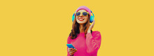 Portrait Of Happy Smiling Modern Young Woman In Wireless Headphones Listening To Music With Smartphone Wearing Knitted Sweater, Pink Hat On Yellow Background