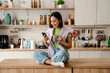 © Drobot Dean - Smiling asian woman using mobile phone and drinking coffee while sitting in kitchen