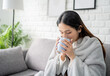 Cold young woman enjoying a mug of hot winter coffee as she wrapped up in blanket in a warm on her sofa at home.