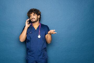 Wall Mural - Good looking Indian male doctor using speaking over or presenting phone, standing isolated over light blue colour. Copy space.