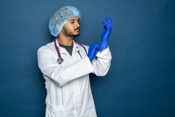 Wall Mural - Indian doctor or physician male looking at hand with focused expression as putting on blue surgical latex glove isolated on blue background